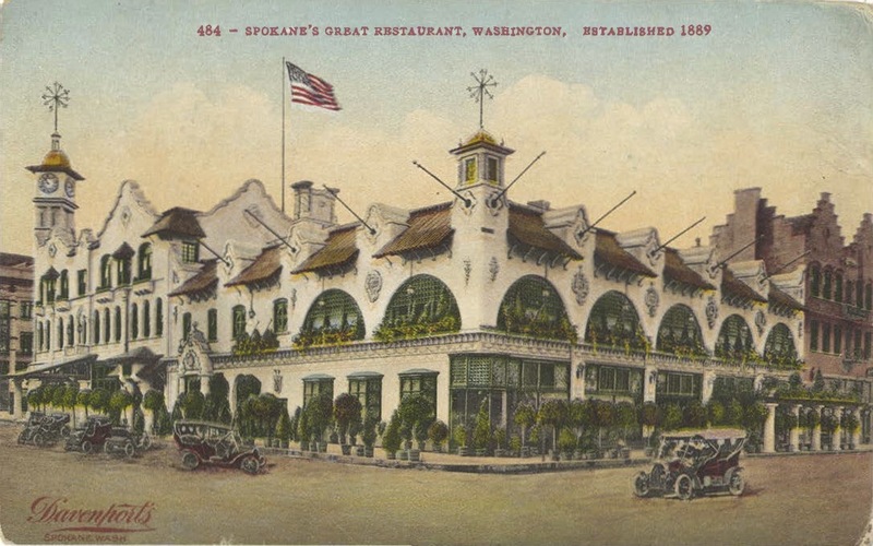 historic colorized postcard depicting a fancy multi-story white building with several early cars parked in front